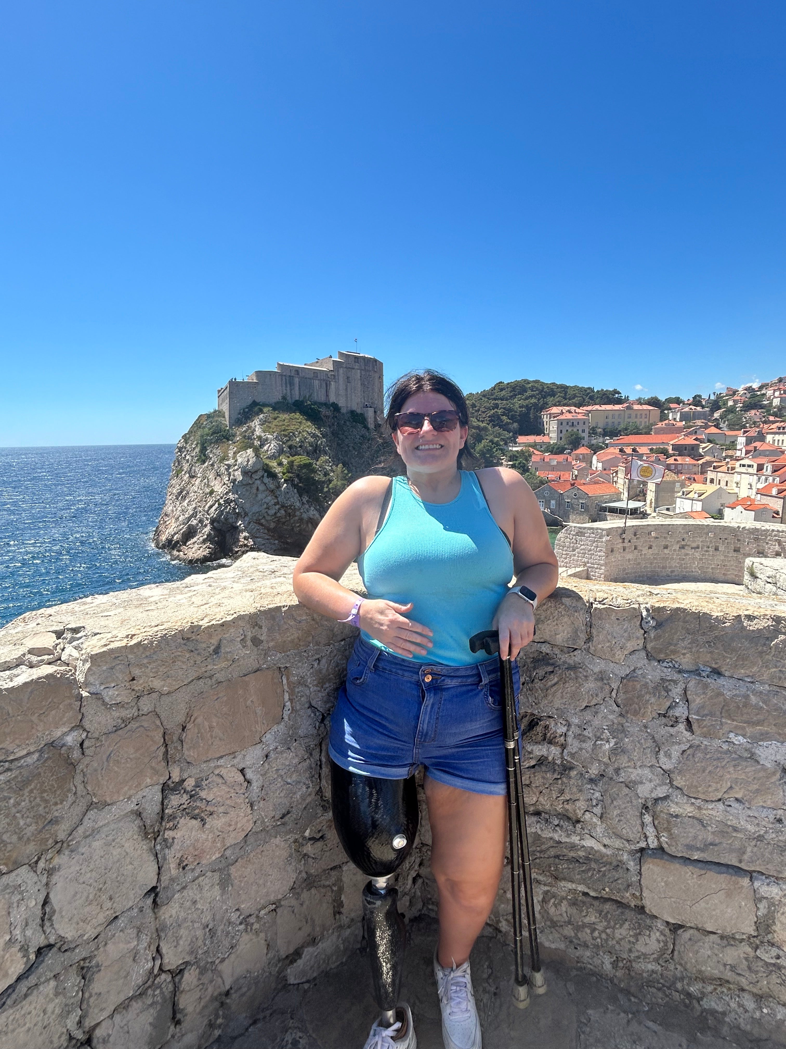 Woman with a prosthetic leg smiling while standing on a stone wall overlooking a coastal town and the sea under a bright blue sky.